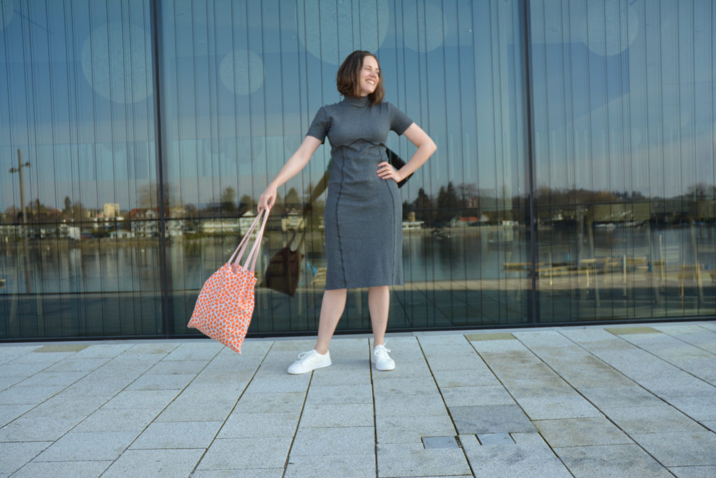 Woman wearing a grey rib knit dress and white sneakers. She is swinging an orange tote back and smiling. Behind her is a large window showing reflections of a lake.