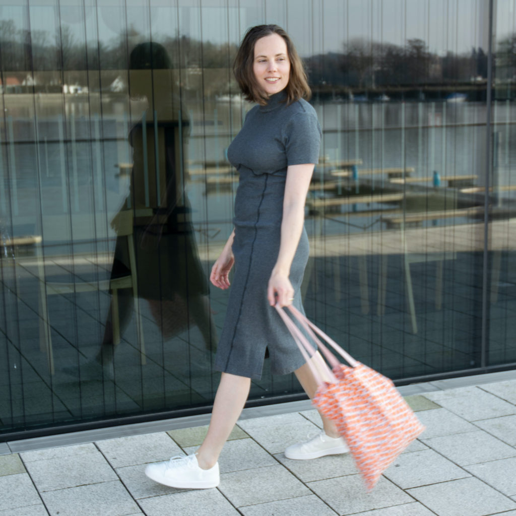 Woman wearing grey dress and white sneakers walking along a large window. She is swinging an orange tote bag and smiling.