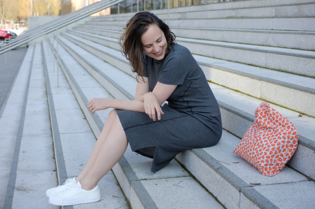 Woman wearing grey dress and white sneakers sitting on a flight of grey stairs. Next to her is an orange tote bag. She is smiling and looking down.