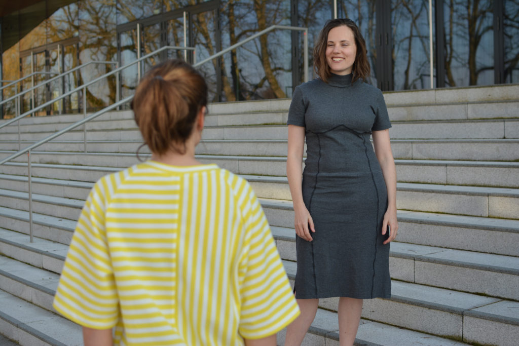 Photograph of a woman wearing a grey dress standing on a flight of stairs. She is smiling and looking toward another woman. The second woman is facing away from the camera, wearing a garment with yellow and white stripes.