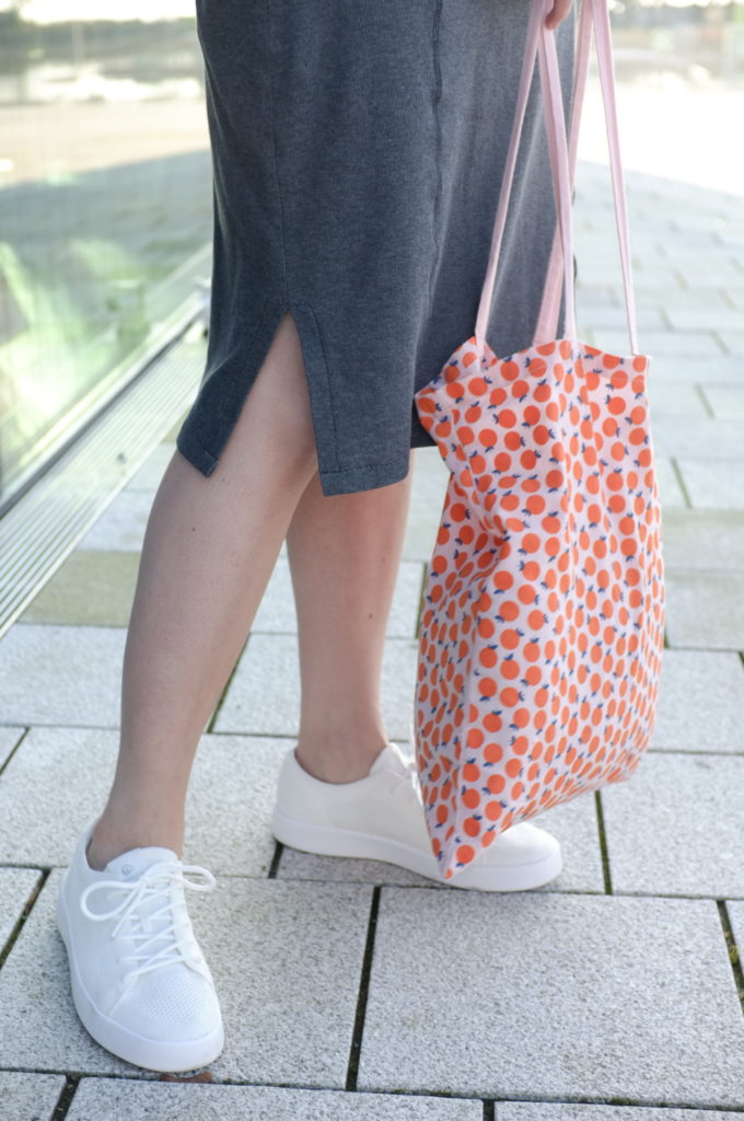 Close-up of the legs of a person wearing a grey dress and white sneakers, holding a tote bag with an orange pattern.