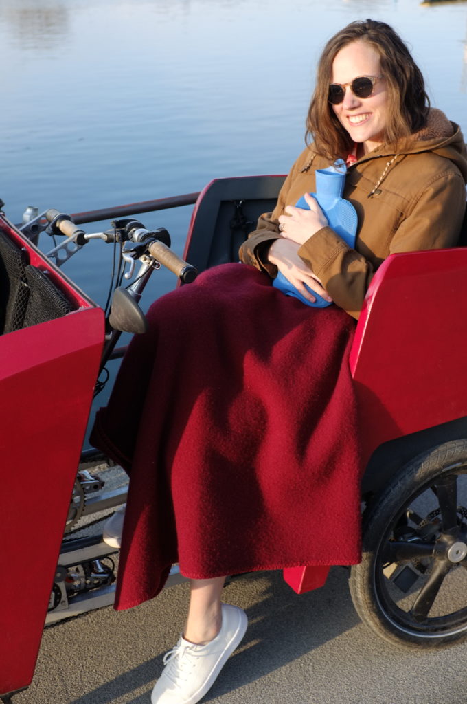 The photograph shows a woman wearing sun glasses sitting in a red vehicle. She is wearing a grey jacket, white sneakers and is wrapped in a red blanket. She is clutching a blue hot-water bottle and smiling. Behind her a body of water is visible.